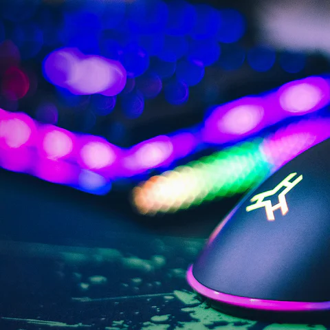 Close-up RGB gaming mouse on a dark desk with colorful keyboard lighting in the background.